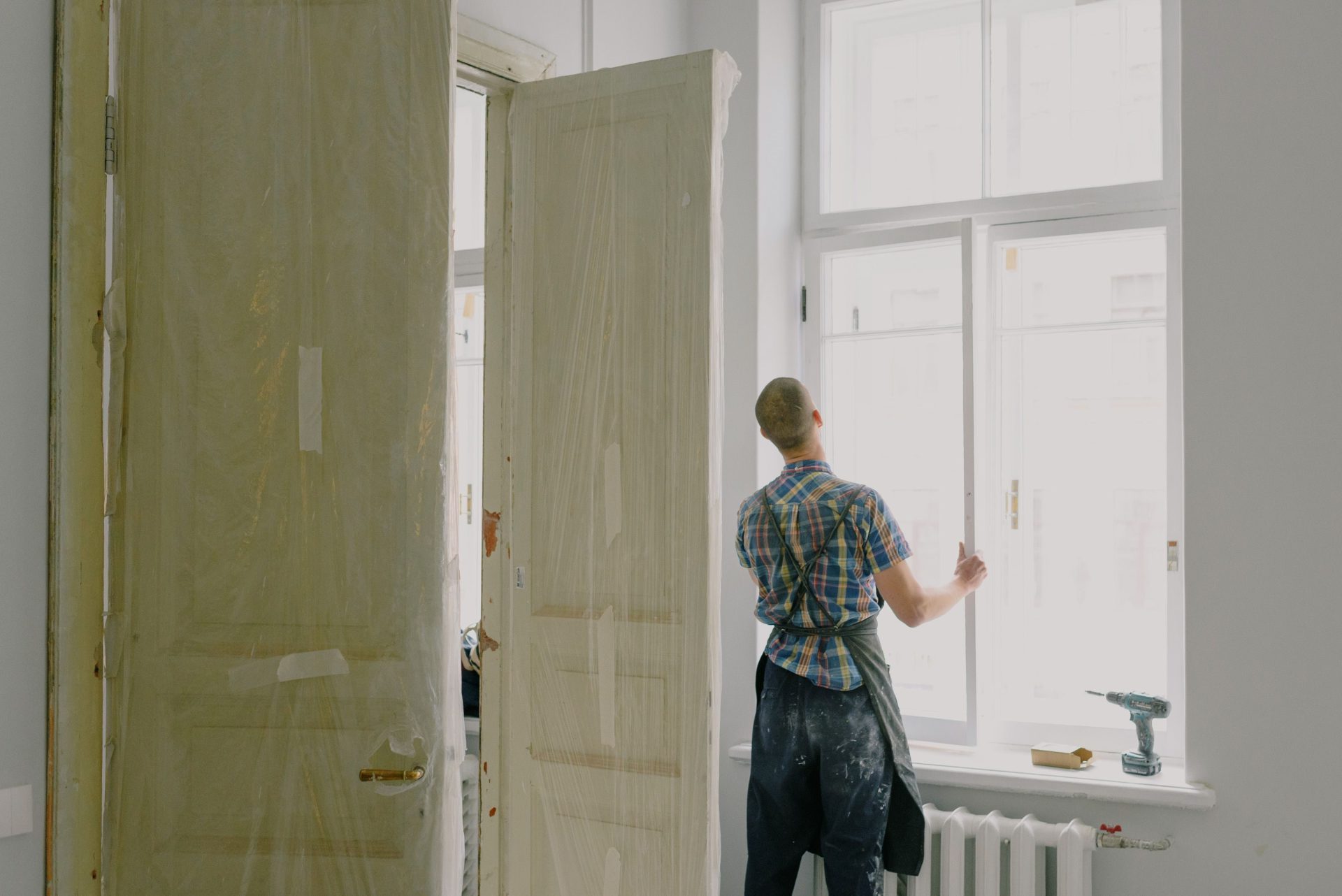 Back view of unrecognizable workman in apron mounting window near drilling machine and doors in flat